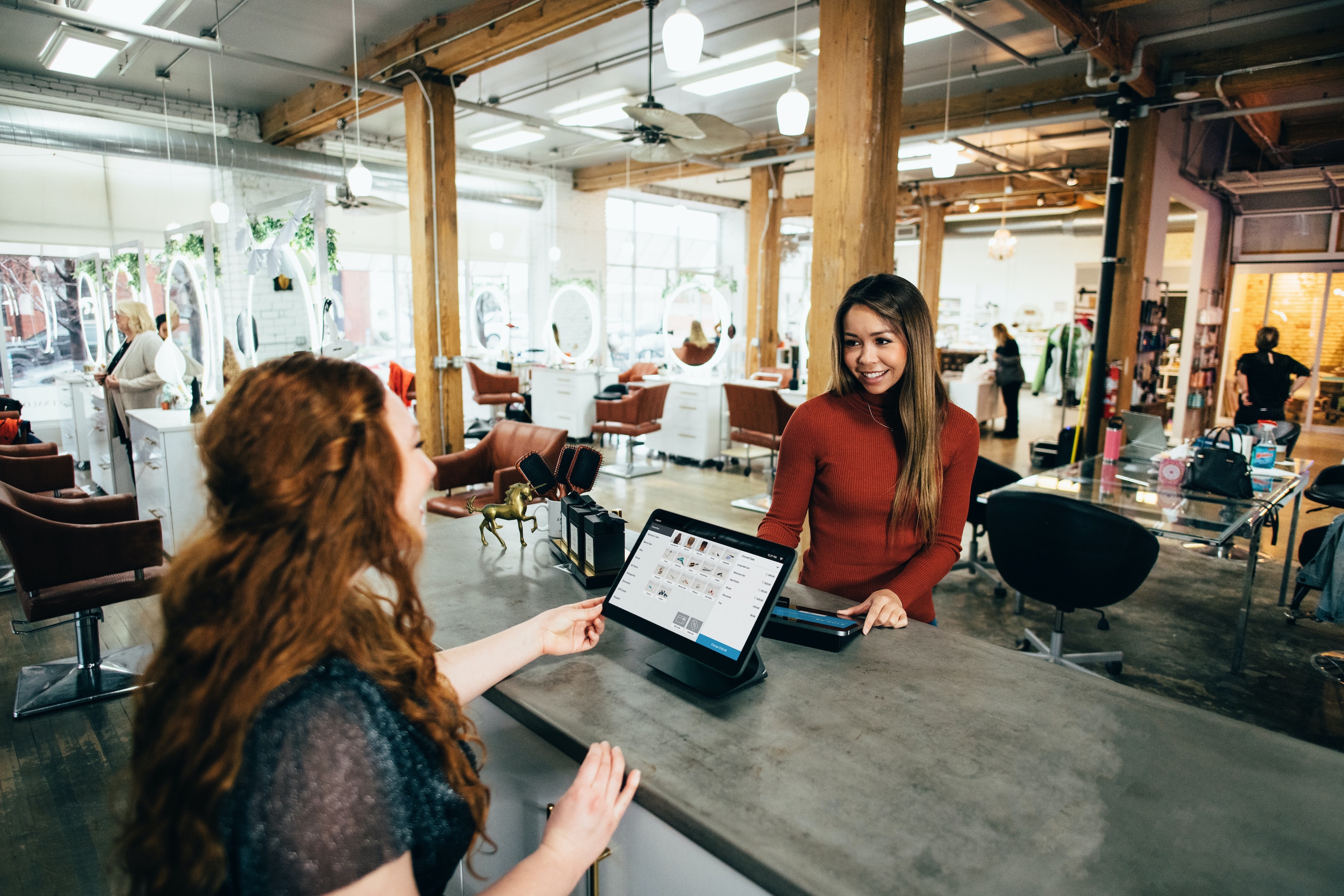 Two women smile at each other across a grey desk, one woman has a mounted tablet and her back faces the viewer